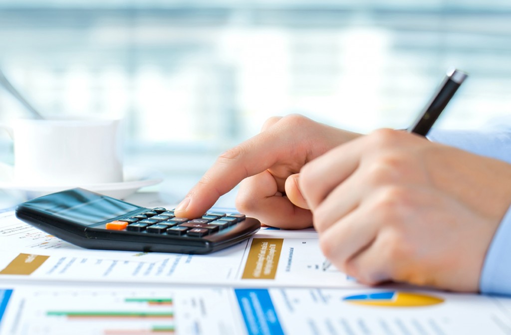 Em uma mesa forrada com relatórios e gráficos, mãos segurando uma caneta e utilizam uma calculadora para fazer o planejamento financeiro da empresa