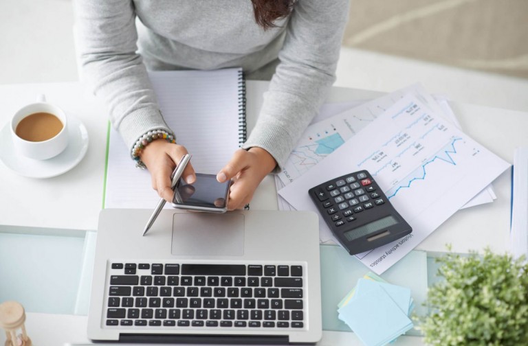 Empreendedora em seu escritório, usando o celular, uma calculadora e um notebook para precificar seus serviços