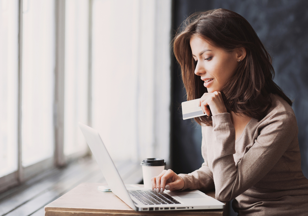 Mulher olhando pensativa enquanto usa um notebook para fazer compras no e-commerce, segurando um cartão de crédito na mão
