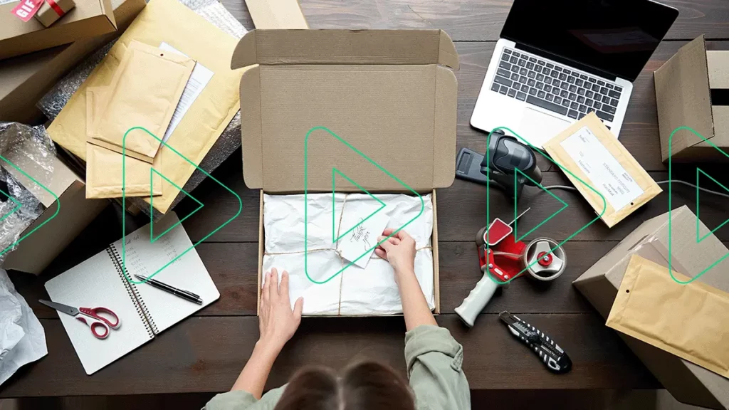 Pessoa empacotando encomenda em cima de uma cheia de utensílios do dia-a-dia de quem trabalha com ecommerce: computador, caderno, tesoura, fita, envelopes, etc.