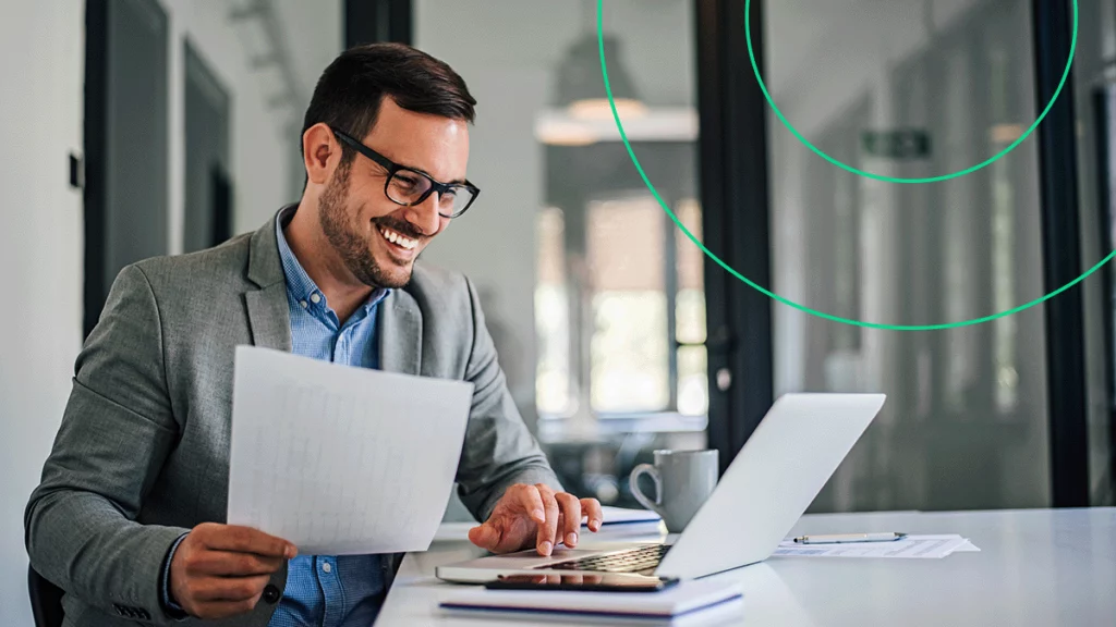 Empresário sorrindo enquanto segura um relatório na mão e digita no notebook, fazendo um Pix com segurança