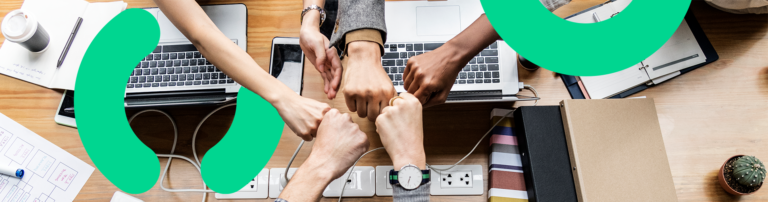 Sobre uma mesa com computadores, cadernos e papéis, várias mãos tocam seus punhos fechados, representando a união entre o Bling e a Locaweb