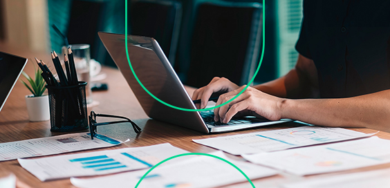 Em uma mesa repleta de relatórios, pessoa trabalha em seu laptop, fazendo o faturamento parcial da empresa